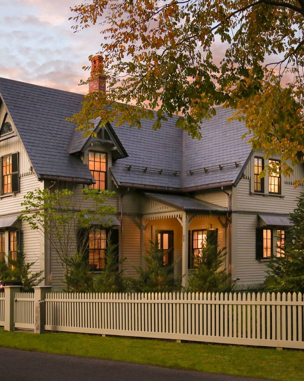 A photo of a house with grey-green painted siding and fence, with stone shingle roofing, antique black gable brackets, and painted colonial detailing.