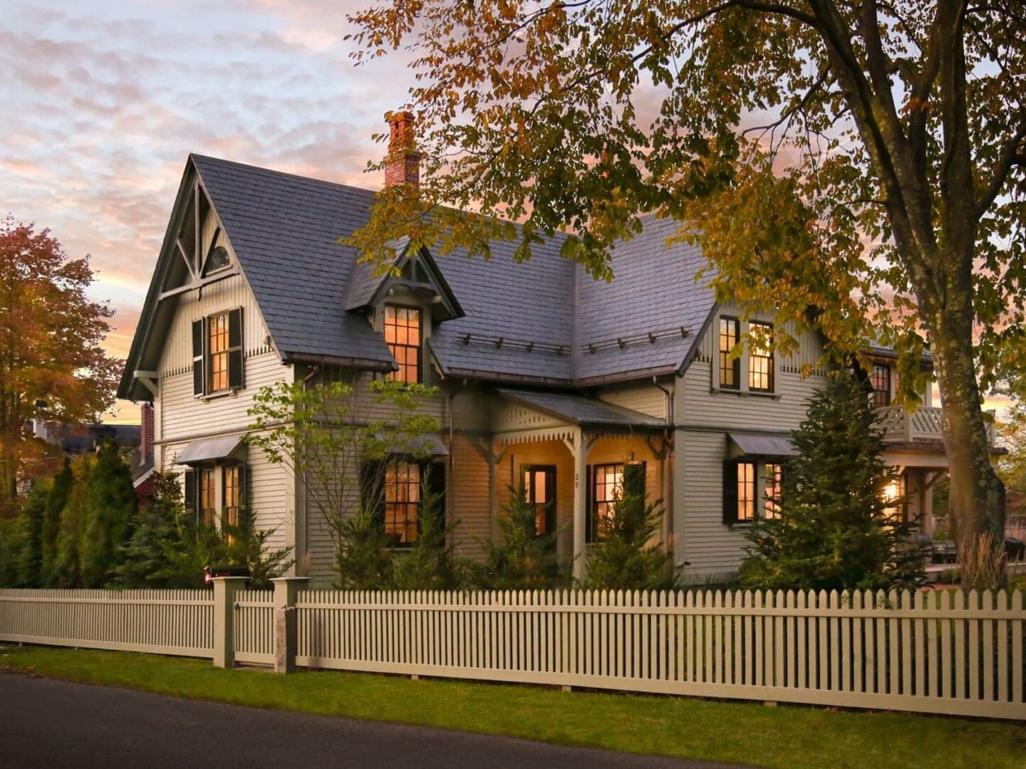 A photo of a house with grey-green painted siding and fence, with stone shingle roofing, antique black gable brackets, and painted colonial detailing.