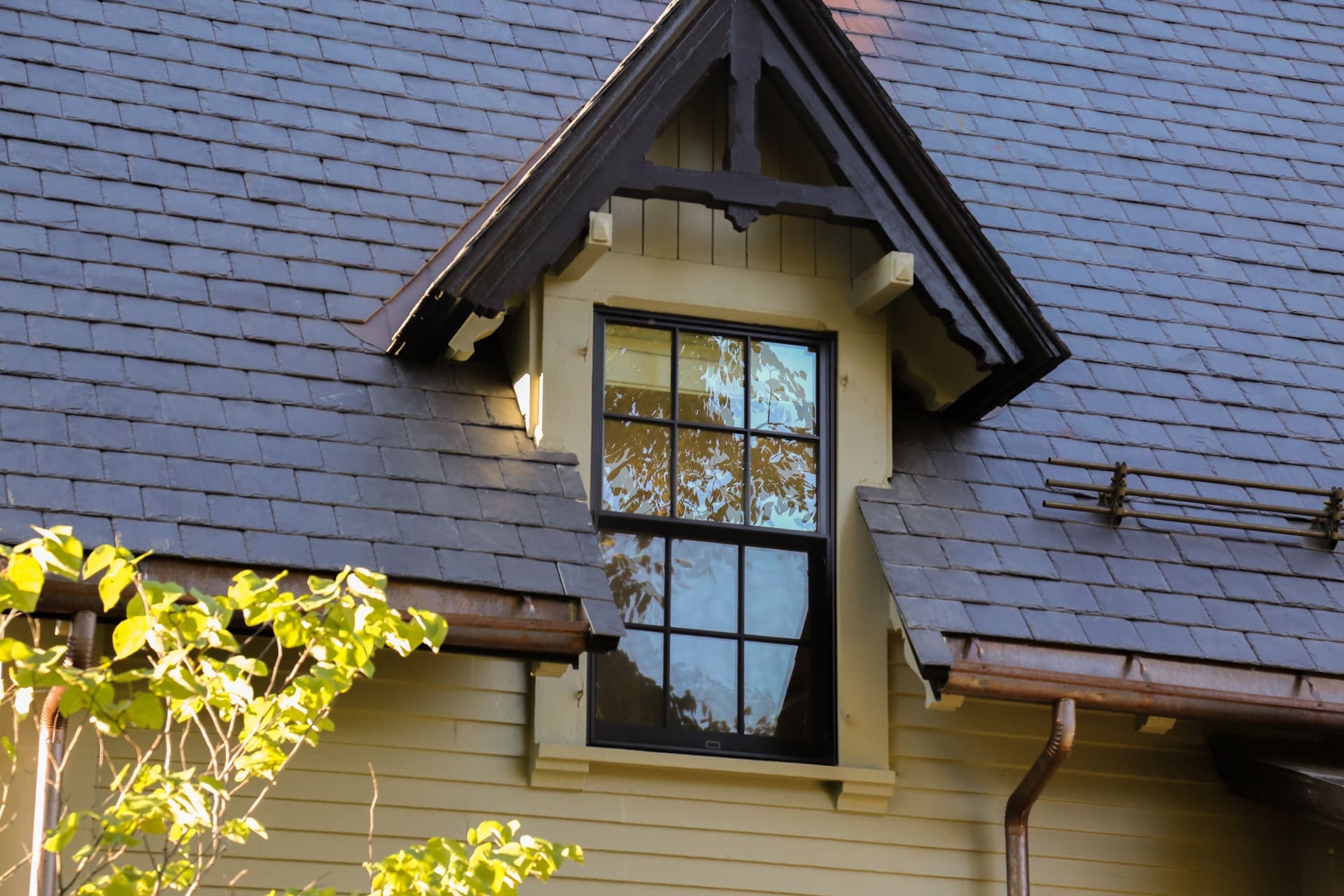 A photo of a grey-green painted house with black stone shingles and a wall dormer window with a painted, black, antique gable bracket.