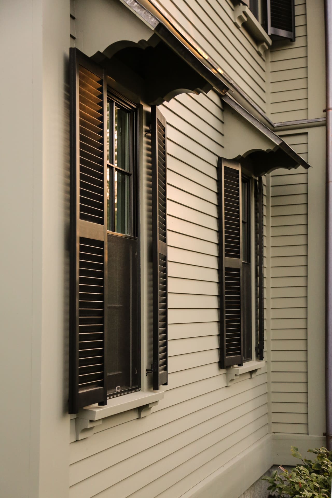 A photo of a house with grey-green siding and windows with black wooden shutters and an overhang.