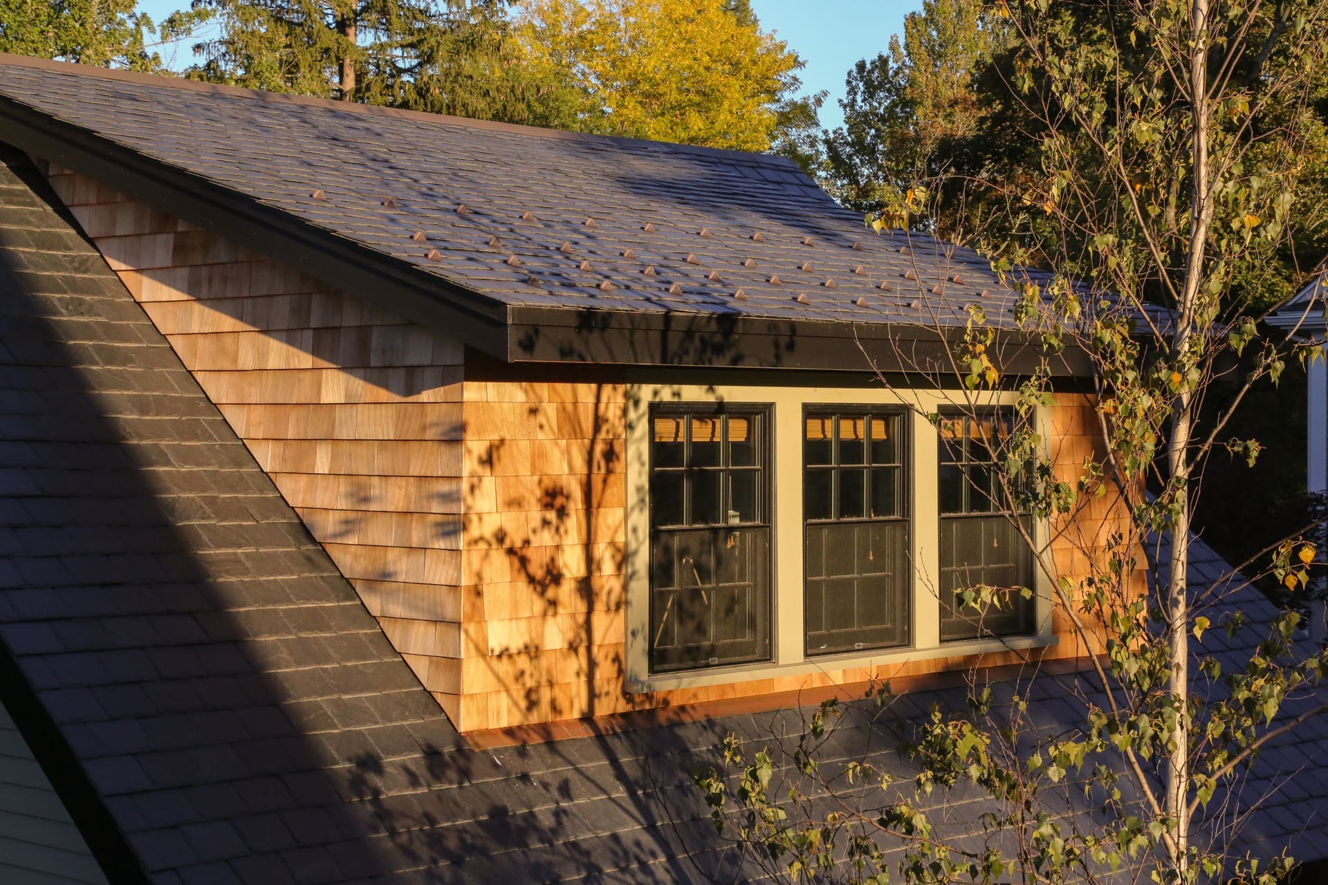 A photo of a dormer roof with black stone shingles, natural wood shingle siding, and grey-green window trim.