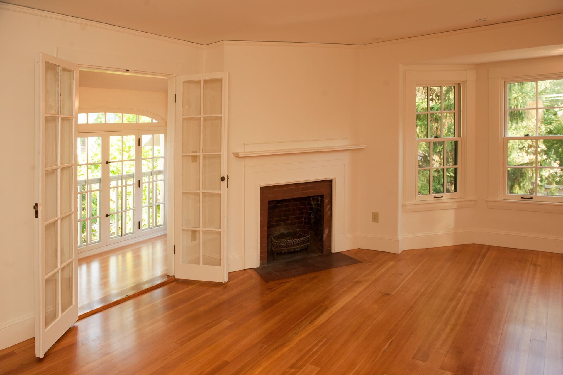 A photo of an empty room with white walls, windows, doors, and hardwood floors, with a dark brick fireplace.