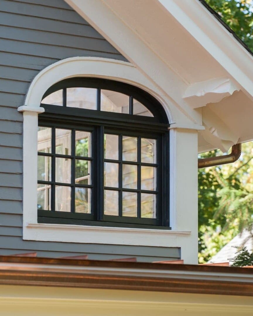 A photo of a black window with curved top and white trim on a house with grey-blue painted siding and a white painted roof overhang.