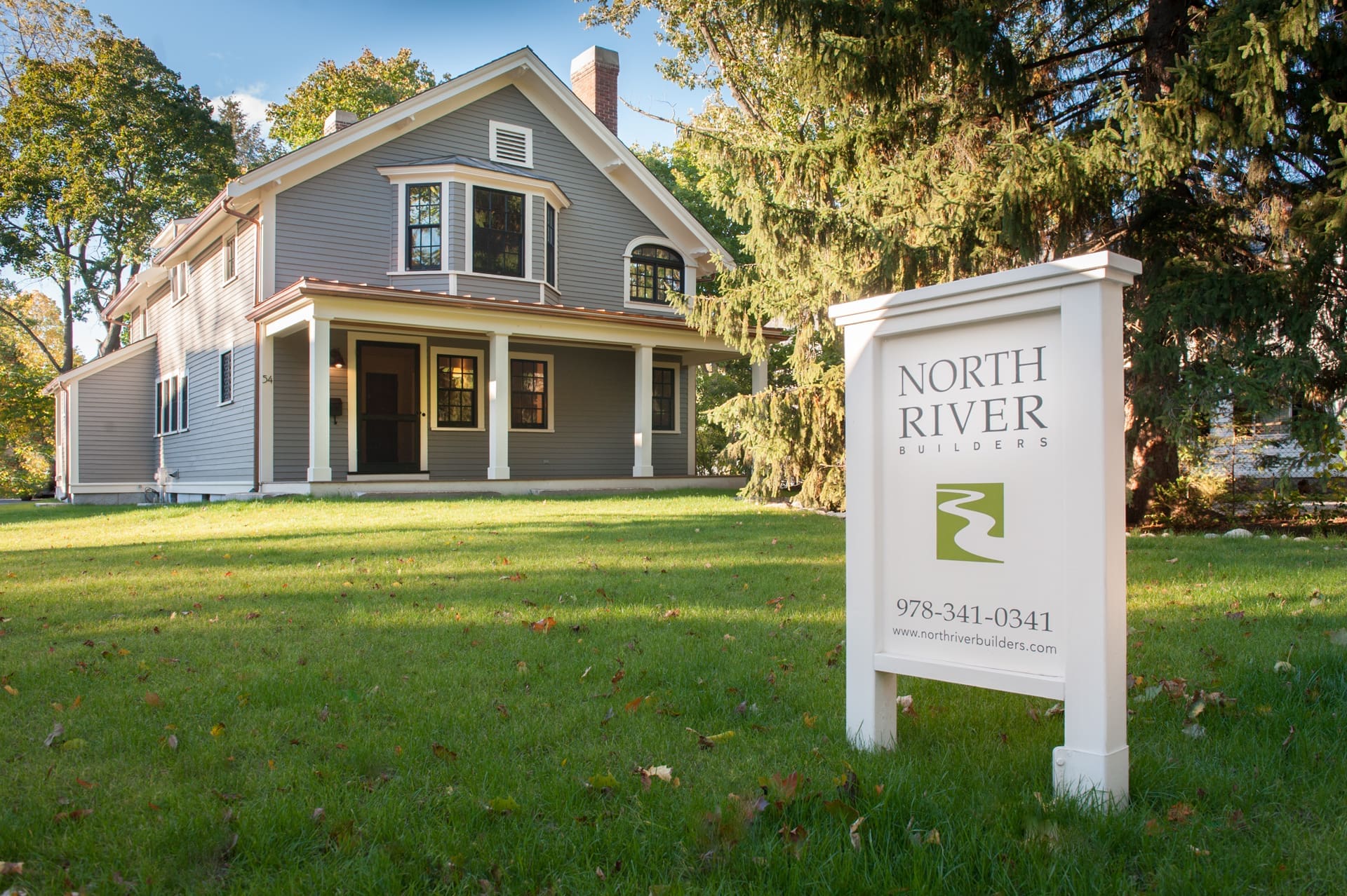 A photo of a North River Builders lawn sign in front of a grey-blue carriage house with front porch overhang, white trim, and black windows.