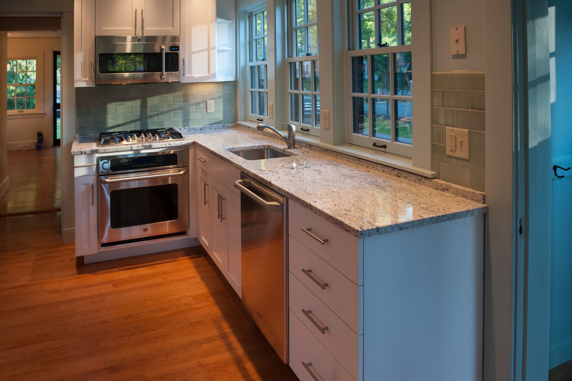 A photo of a kitchenette with white cabinets, stainless steel appliances, grey stone countertops, and a bank of colonial-style windows.