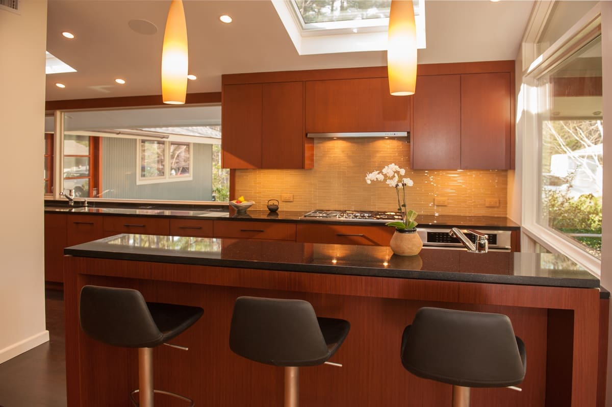 A photo of a mid-century modern kitchen with three barstools, cherry wooden cabinetry, deck house-style windows, and black stone countertops.