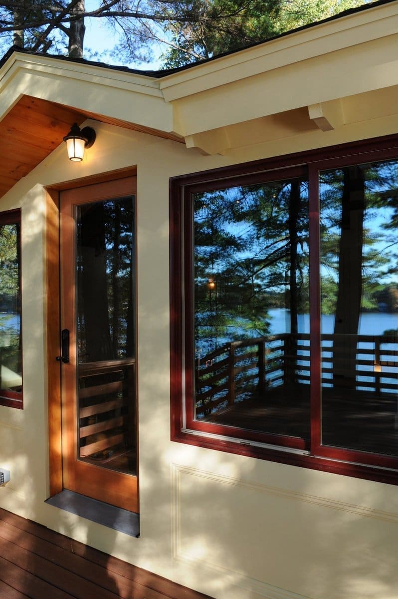 A photo of a front porch with cherry wooden windows and a glass door with a roof gable above it, surrounded by forest and water.