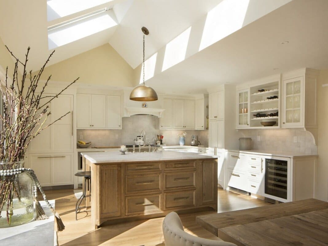 A photo of a kitchen with white cabinets, stainless steel appliances, an island made of weathered wood, white marble countertops, and vaulted ceilings with skylights.