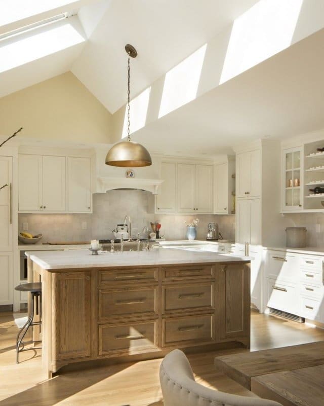 A photo of a kitchen with white cabinets, stainless steel appliances, an island made of weathered wood, white marble countertops, and vaulted ceilings with skylights.
