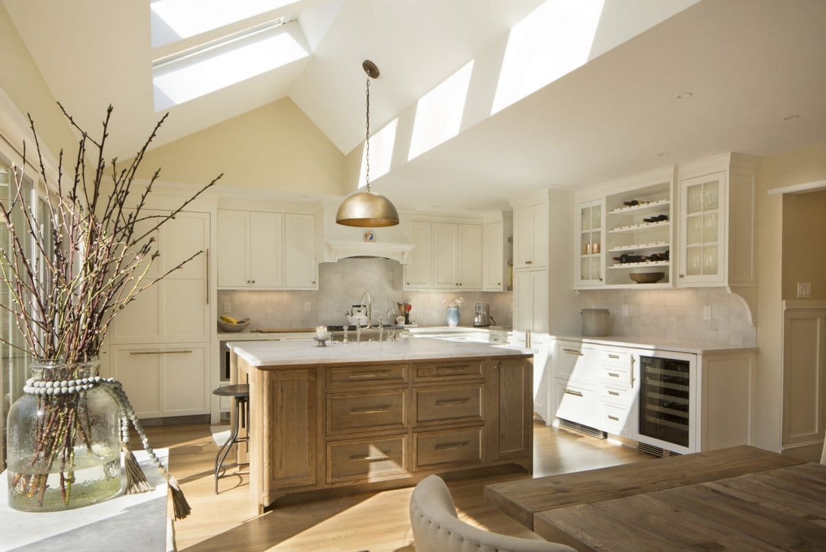 A photo of a kitchen with white cabinets, stainless steel appliances, an island made of weathered wood, white marble countertops, and vaulted ceilings with skylights.