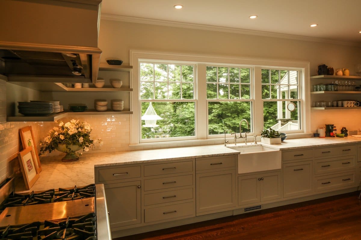 A photo of a kitchen with shaker-style cabinets, a deep porcelain sink, overhead shelving storage, large windows, stainless steel appliances, and hardwood floors.