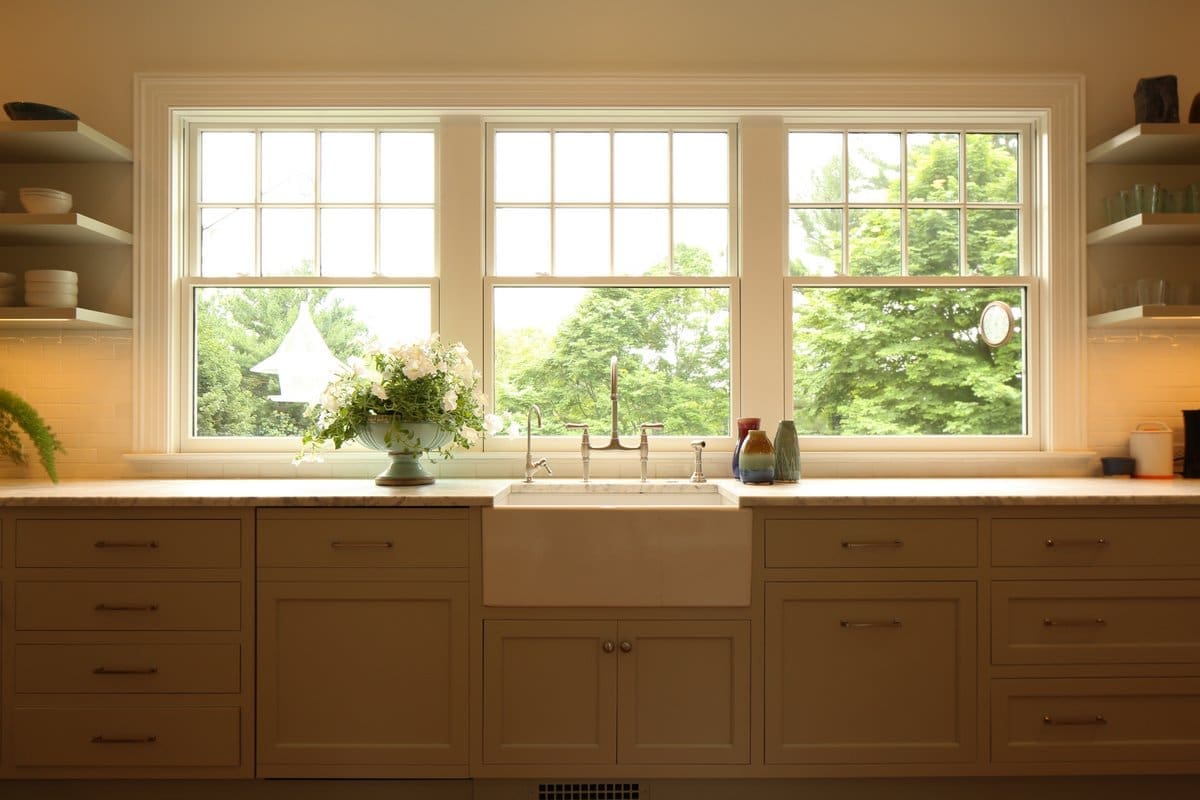 A photo of a kitchen with shaker-style cabinets, a large porcelain sink, and three windows with a view to the forest.