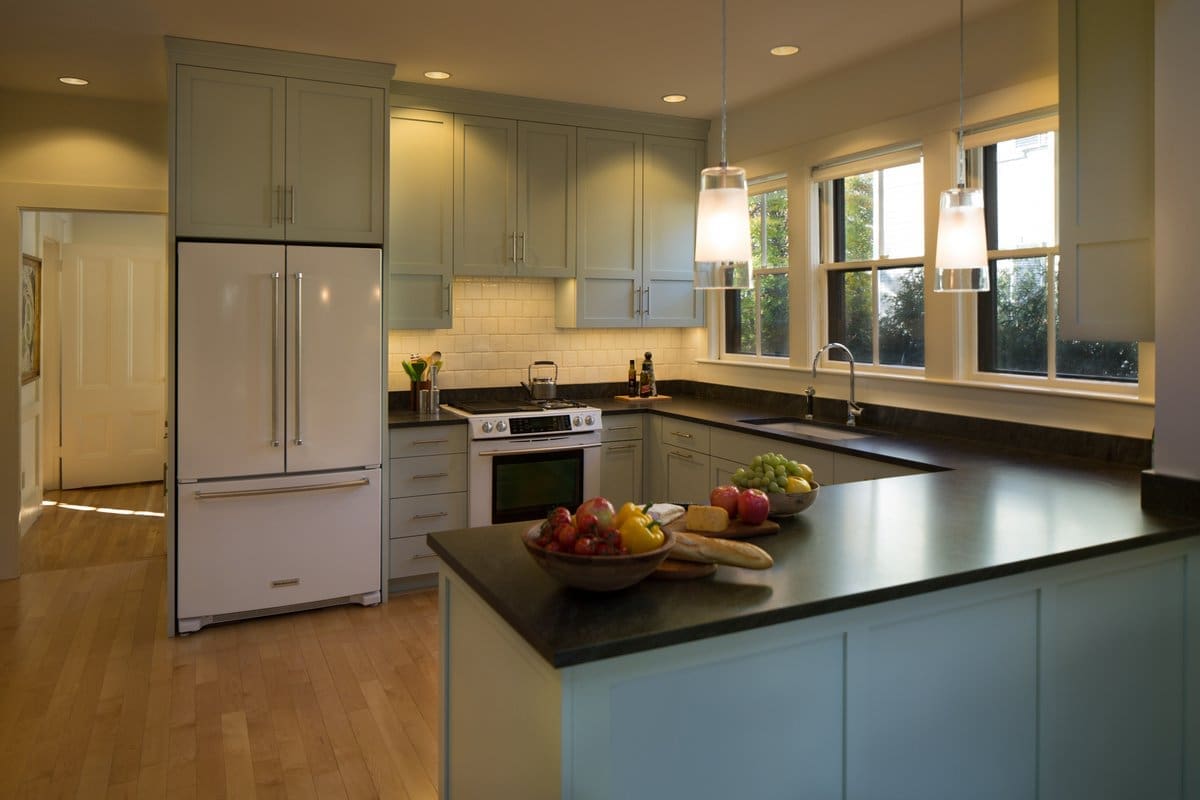 A photo of a kitchen with black stone countertops, faded green painted shaker-style cabinets, and white appliances.