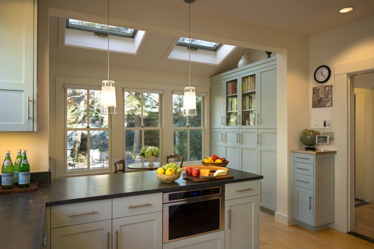 A photo of a kitchen and dining room with faded light green painted shaker-style cabinets and built-ins, skylights, black stone countertops, and large windows.