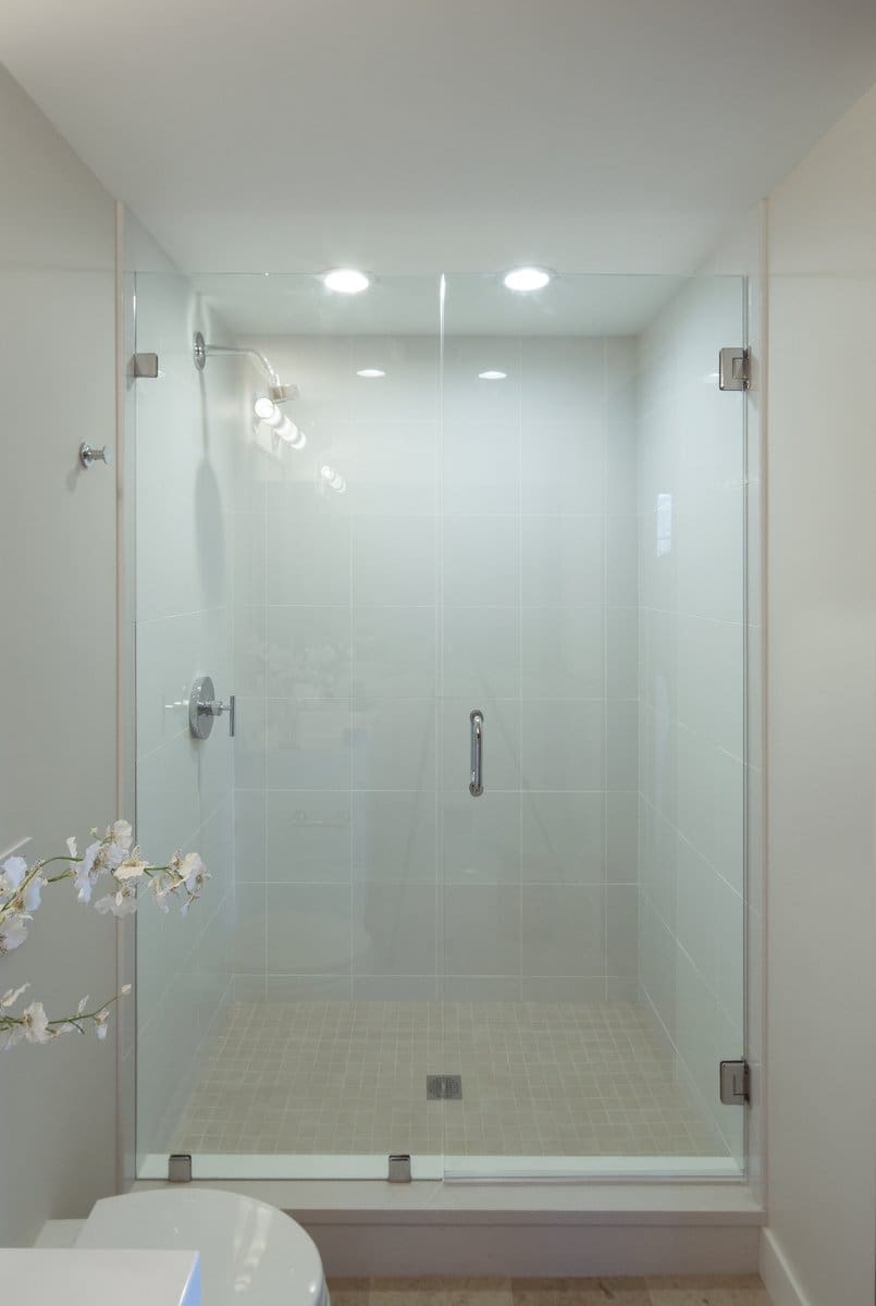 A photo of a shower with light blue square tiled walls, light beige small square tiled floor, and glass doors.