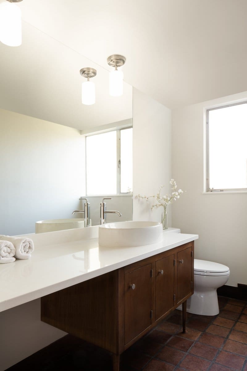 A photo of a bathroom with white walls, a white marble vanity and wooden cabinet, silver hardware, and square stone floor tile.