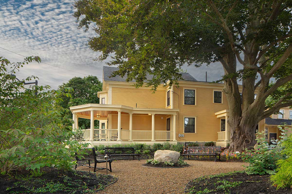 A photo of a yellow building with a front porch with yellow columns and railings and a garden.