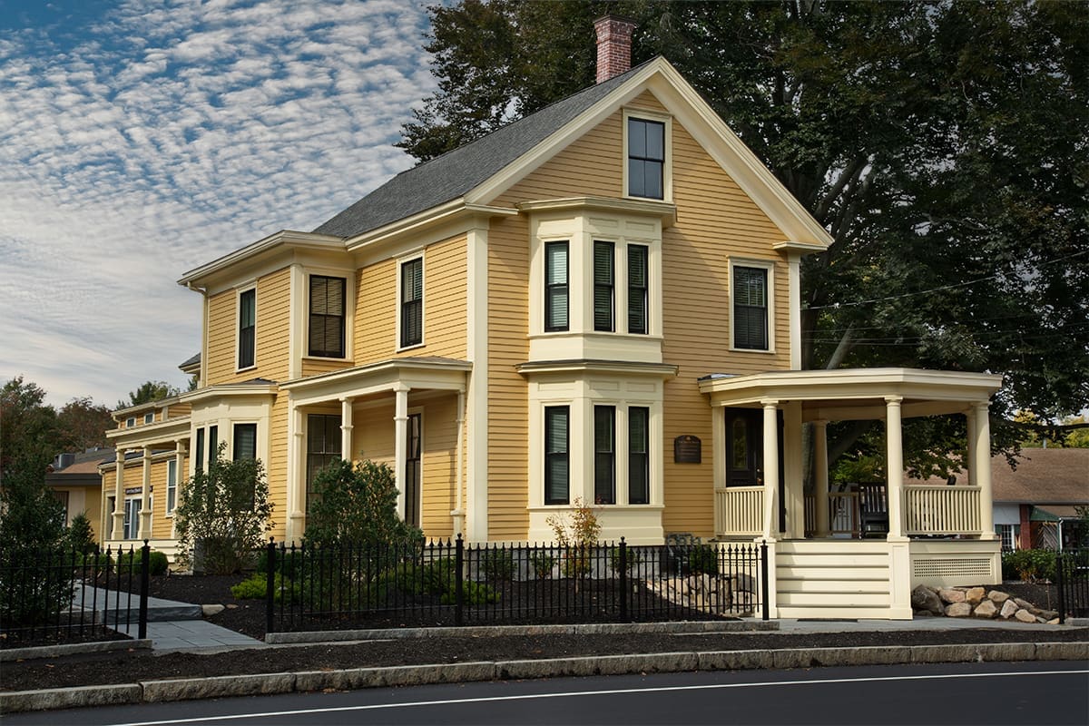 A photo of a yellow building with a front porch with yellow columns and railings and a black fence from street view.
