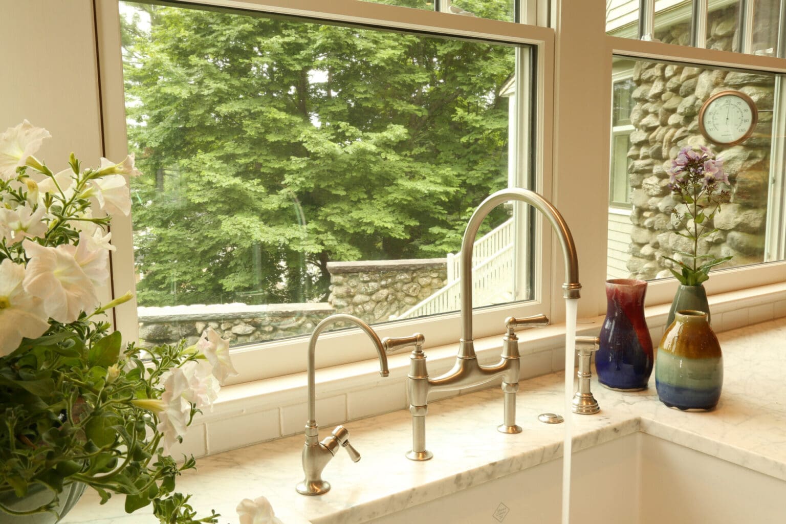 A photo of a running faucet in front of a large window in a kitchen with marble countertops.