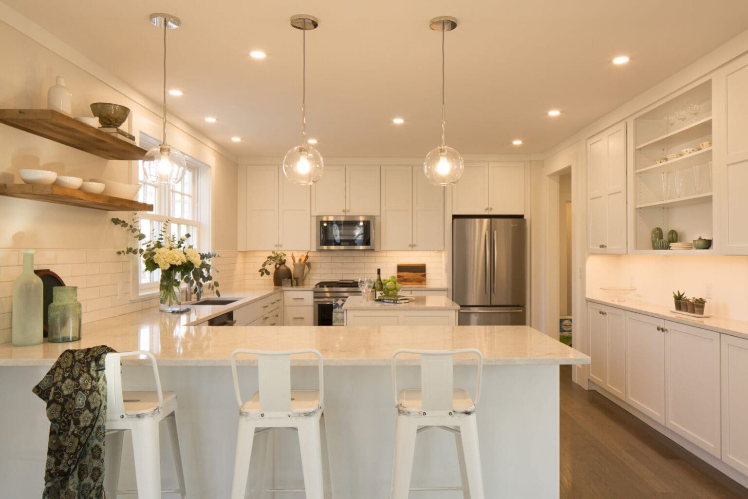 A photo of a kitchen with white painted shaker-style cabinets, three white counter-side chairs, stainless steel appliances, white subway tile backsplash, wooden shelves, light grey marble countertops, and an island.