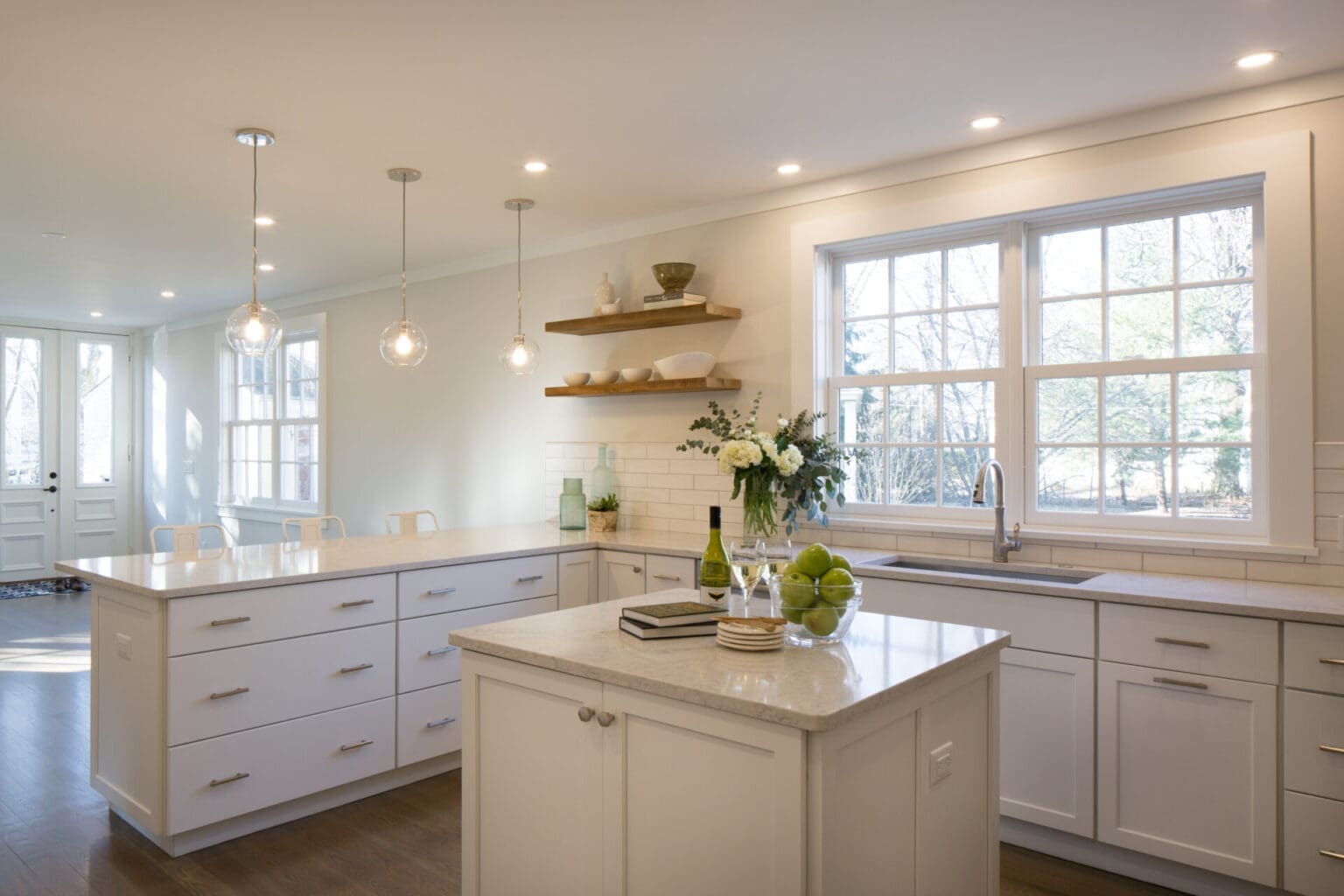 A photo of a kitchen with white painted shaker-style cabinets, stainless steel appliances, white subway tile backsplash, wooden shelves, light grey marble countertops, an island, and three white chairs.