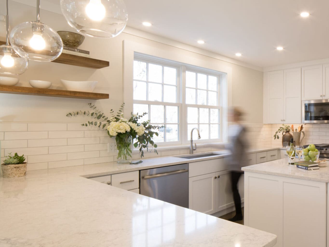 A photo of a kitchen with white painted shaker-style cabinets, stainless steel appliances, white subway tile backsplash, wooden shelves, and light grey marble countertops.