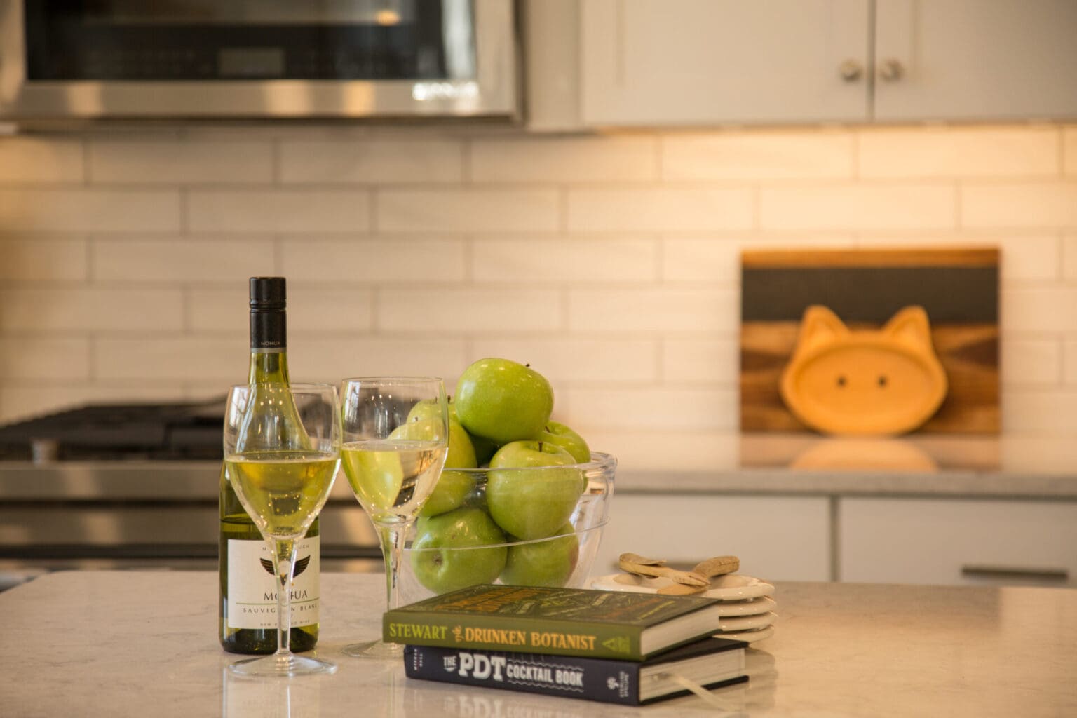 A photo of green apples in a bowl, cookbooks, and two glasses and a bottle of white wine in a kitchen with grey marble countertops.