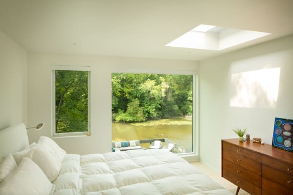 A photo of a bedroom with windows looking out onto a lake, a skylight, and a wooden dresser.