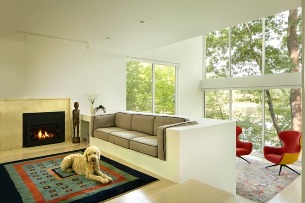 A picture of a living room with a dog sitting on a colorful rug, a couch built into a staircase, a black fireplace, and floor-to-ceiling windows.