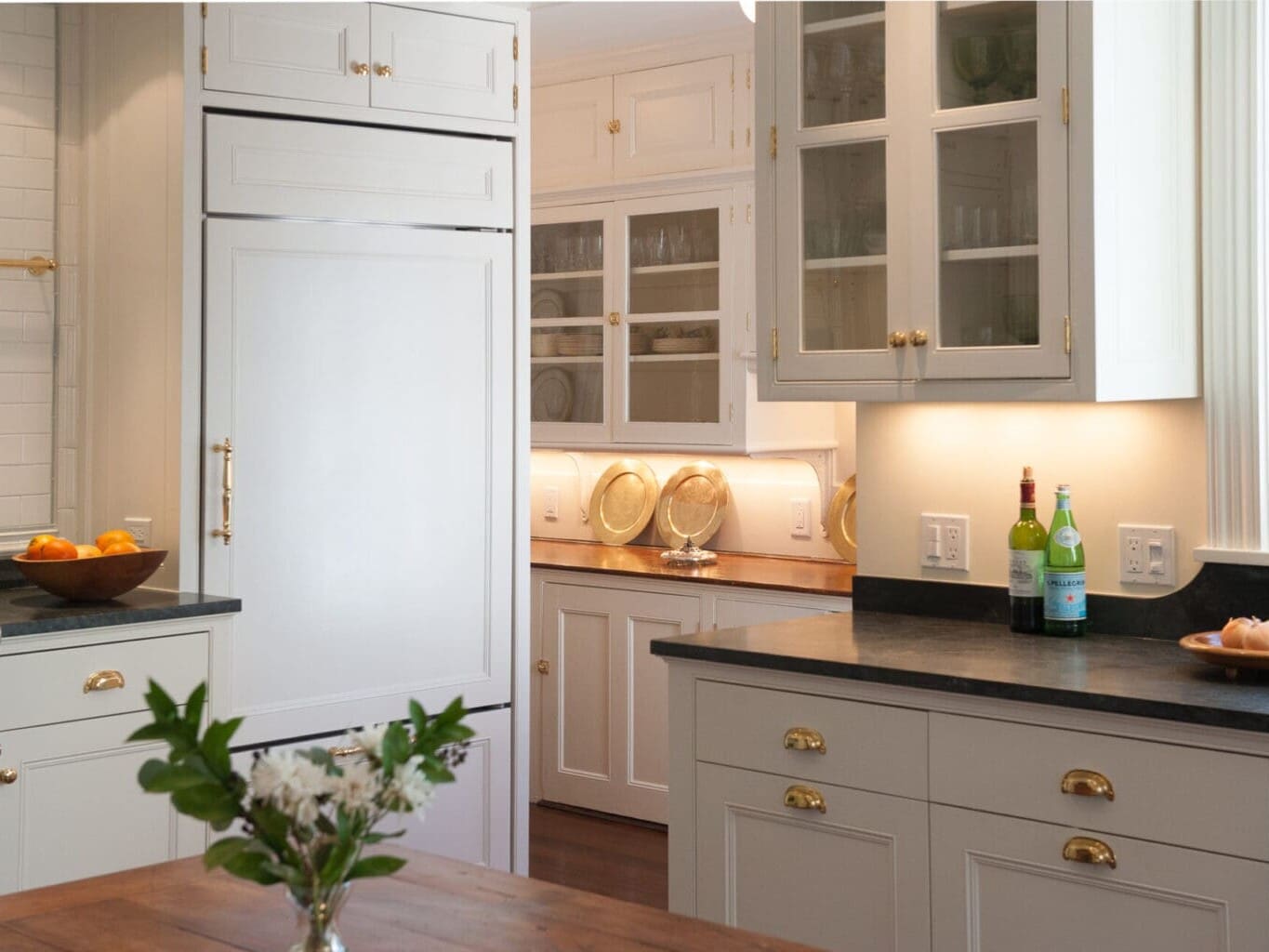 A photo of a kitchen with painted shaker-style cabinetry, brass hardware, a built-in fridge with shaker-style paneling, and black marble countertops.