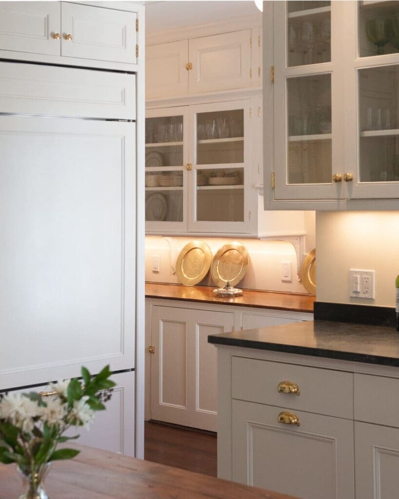 A photo of a kitchen with painted shaker-style cabinetry, brass hardware, a built-in fridge with shaker-style paneling, and black marble countertops.