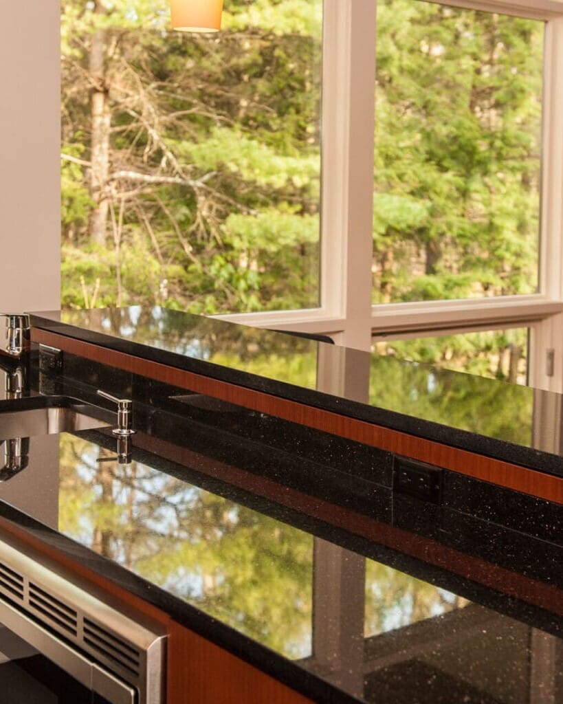 A photo of a mid-century modern kitchen island with black stone countertops, stainless steel appliances, black hardware, and floor-to-ceiling windows.