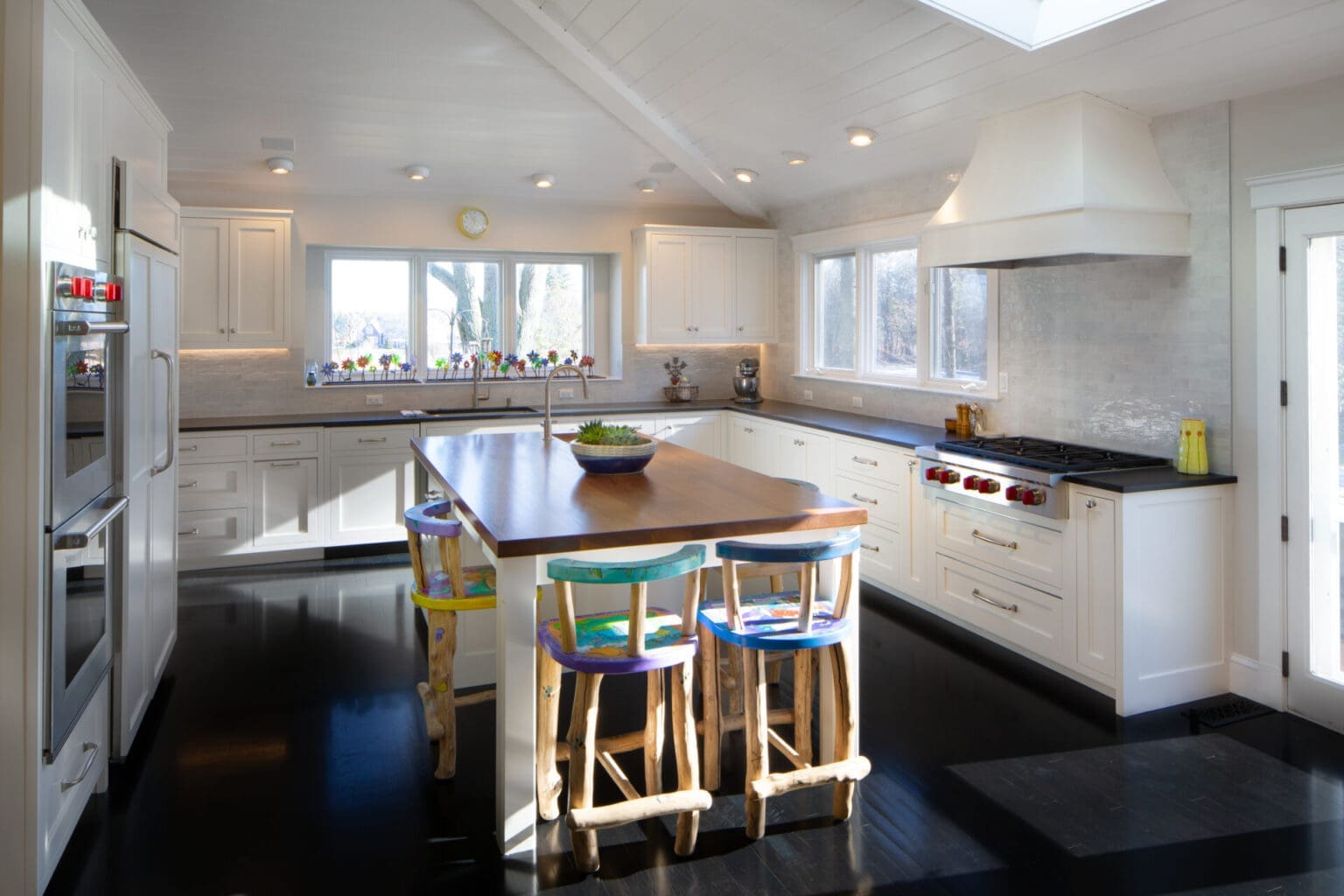 A photo of a large kitchen with white shaker-style cabinetry and black countertops. An island with a wood countertop and a secondary sink is in the middle of the kitchen, surrounded by four brightly painted wooden chairs.