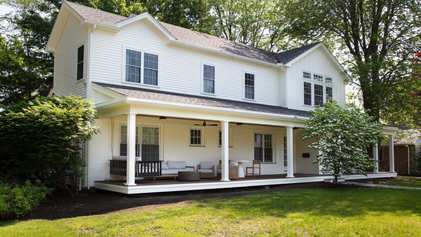 A photo of a white two-level house with a farmers porch with white square pillars, dark brown wooden flooring, and assorted outdoor furniture with grey cushions.