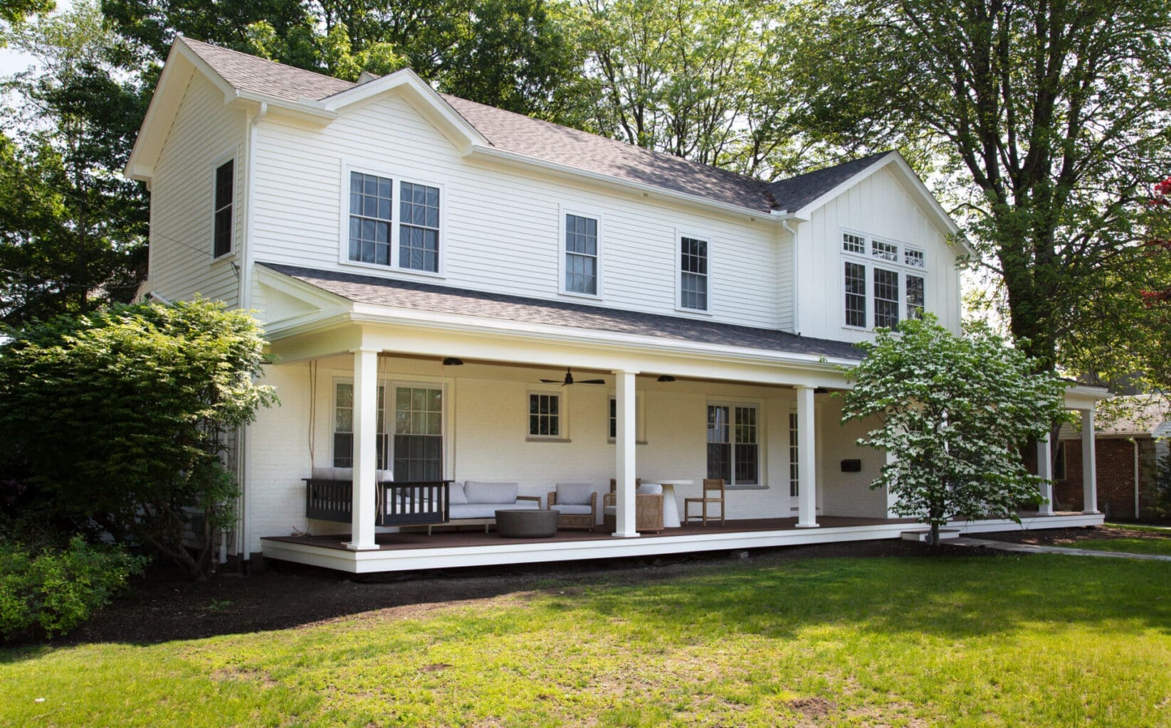 A photo of a white two-level house with a farmers porch with white square pillars, dark brown wooden flooring, and assorted outdoor furniture with grey cushions.