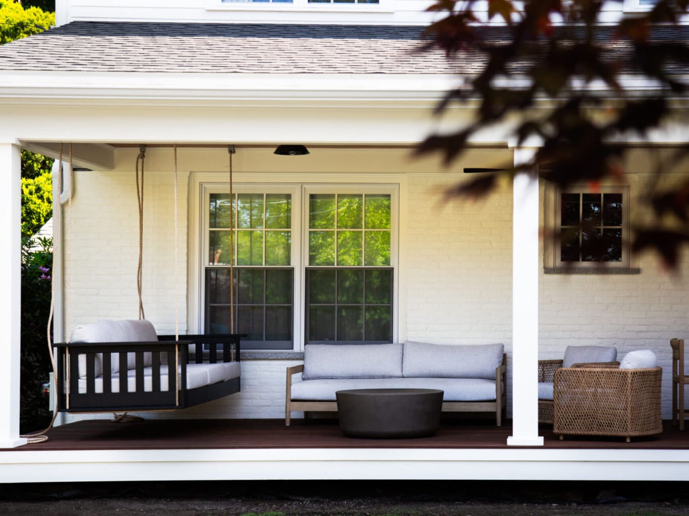 A photo of a farmers porch with white square pillars, white brick wall, dark brown wooden flooring, and assorted outdoor furniture with grey cushions.