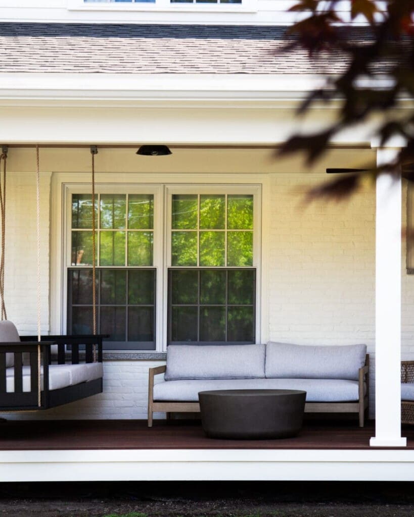 A photo of a farmers porch with white square pillars, white brick wall, dark brown wooden flooring, and assorted outdoor furniture with grey cushions.