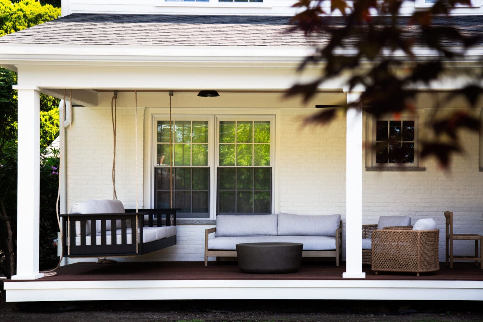 A photo of a farmers porch with white square pillars, white brick wall, dark brown wooden flooring, and assorted outdoor furniture with grey cushions.