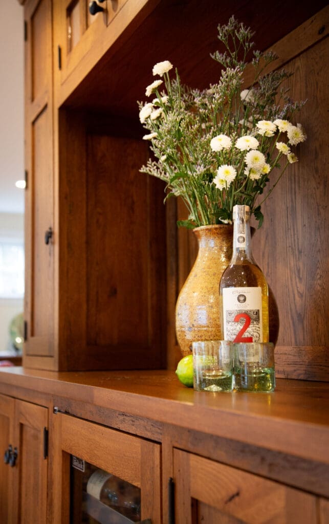 A photo of a golden vase, tequila bottle, a lime, and two glasses on a wooden cabinet bar.