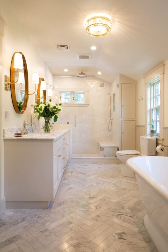 A photo of a bathroom with fishbone-tiled marble floors, white shaker-style cabinetry with brass hardware, marble counter, two sinks and mirrors, and a shower with glass door.