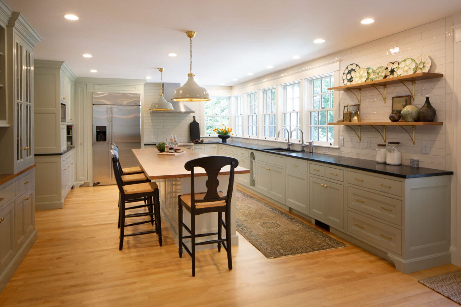 Photo of a classic grey-teal kitchen with shaker-style cabinets, brass hardware, stone countertops, a bank of windows, and a large island with natural wooden top.