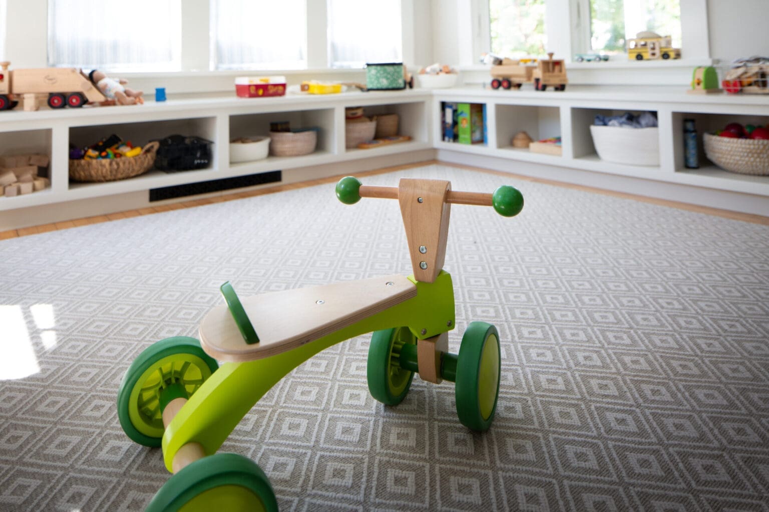 A photo of a green toy bicycle in a child's playroom with built-in storage and large windows.