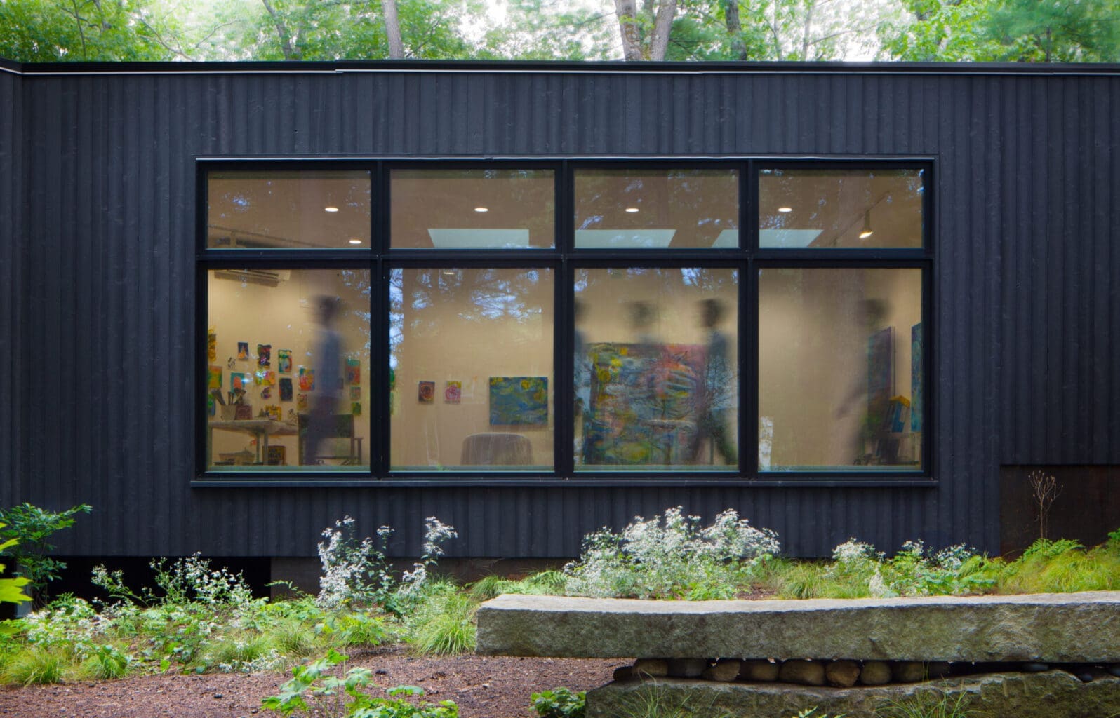 A photo of the outside of the gallery built from Japanese 'shou sugi ban' wood. Four windows show colorful paintings inside.