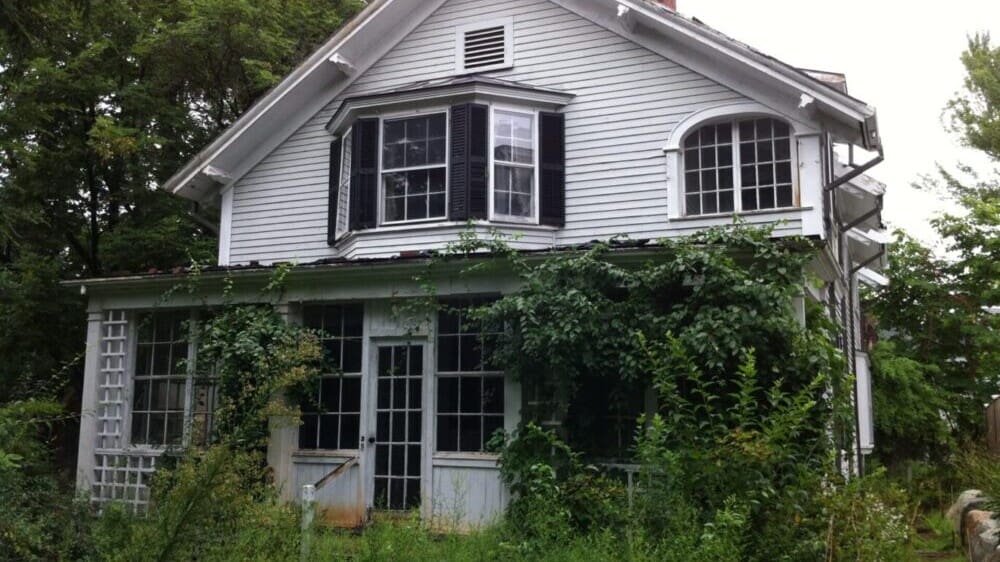 Photo of a chipped-white colonial house covered in overgrown plants.