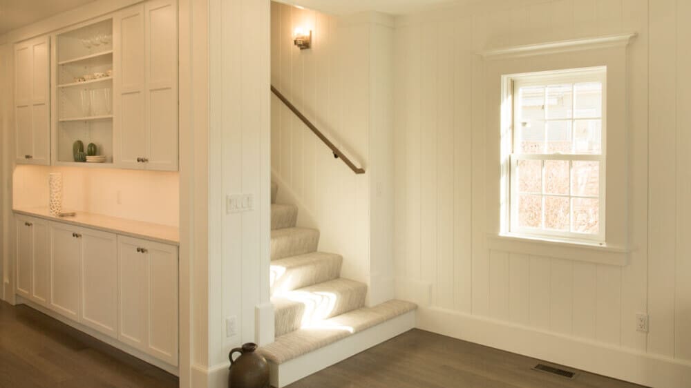 A photo of a bright, finished, white room with built-in cabinetry, refurbished windows, re-sided walls, new flooring, and a renovated stairway.