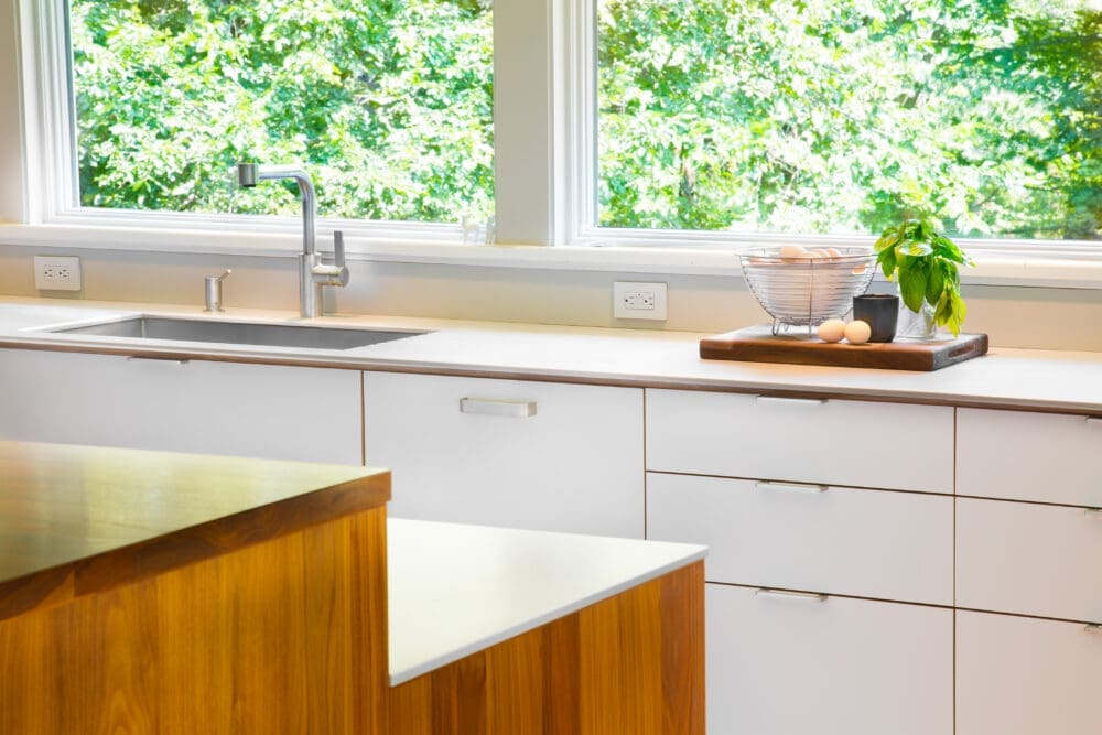 A photo of a white mid-century modern-style kitchen with large windows, a stainless steel sink, and an island.