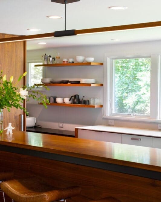 A photo of a bright mid-century modern-style kitchen with a white countertop and cabinets, a stainless steel sink, wooden shelving, and a cherry wooden island