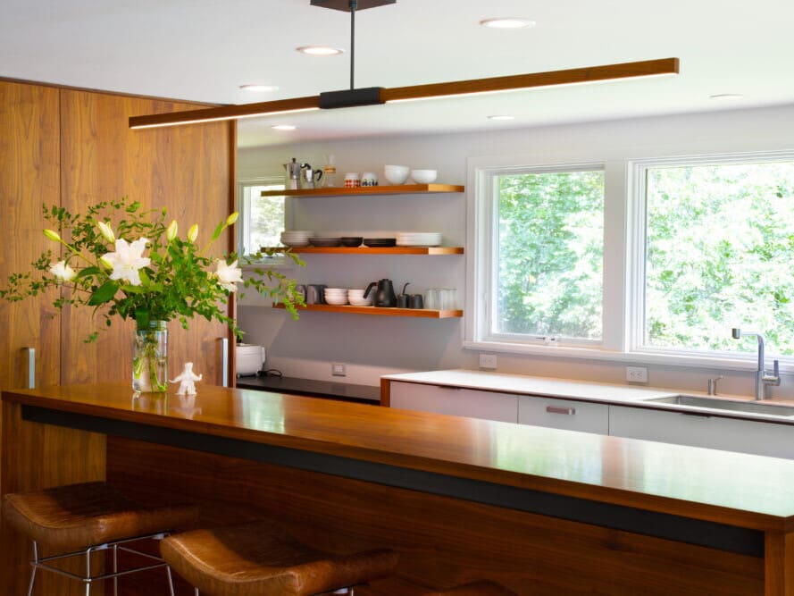 A photo of a bright mid-century modern-style kitchen with a white countertop and cabinets, a stainless steel sink, wooden shelving, and a cherry wooden island
