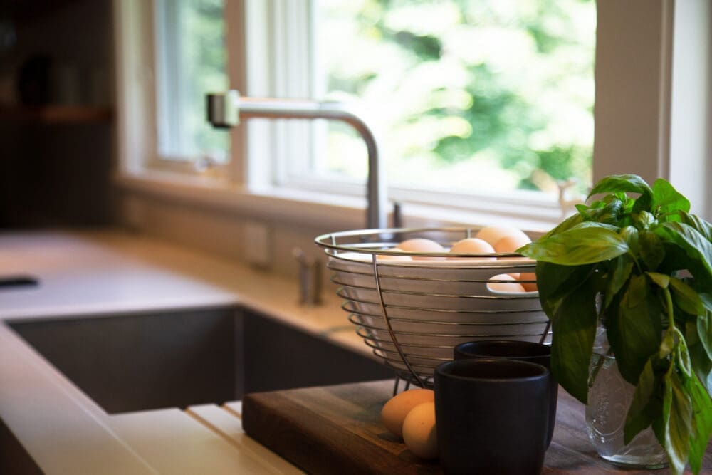 A photo of a white marble kitchen counter with large windows and a stainless steel sink, a basket of eggs, two mugs, and a plant.