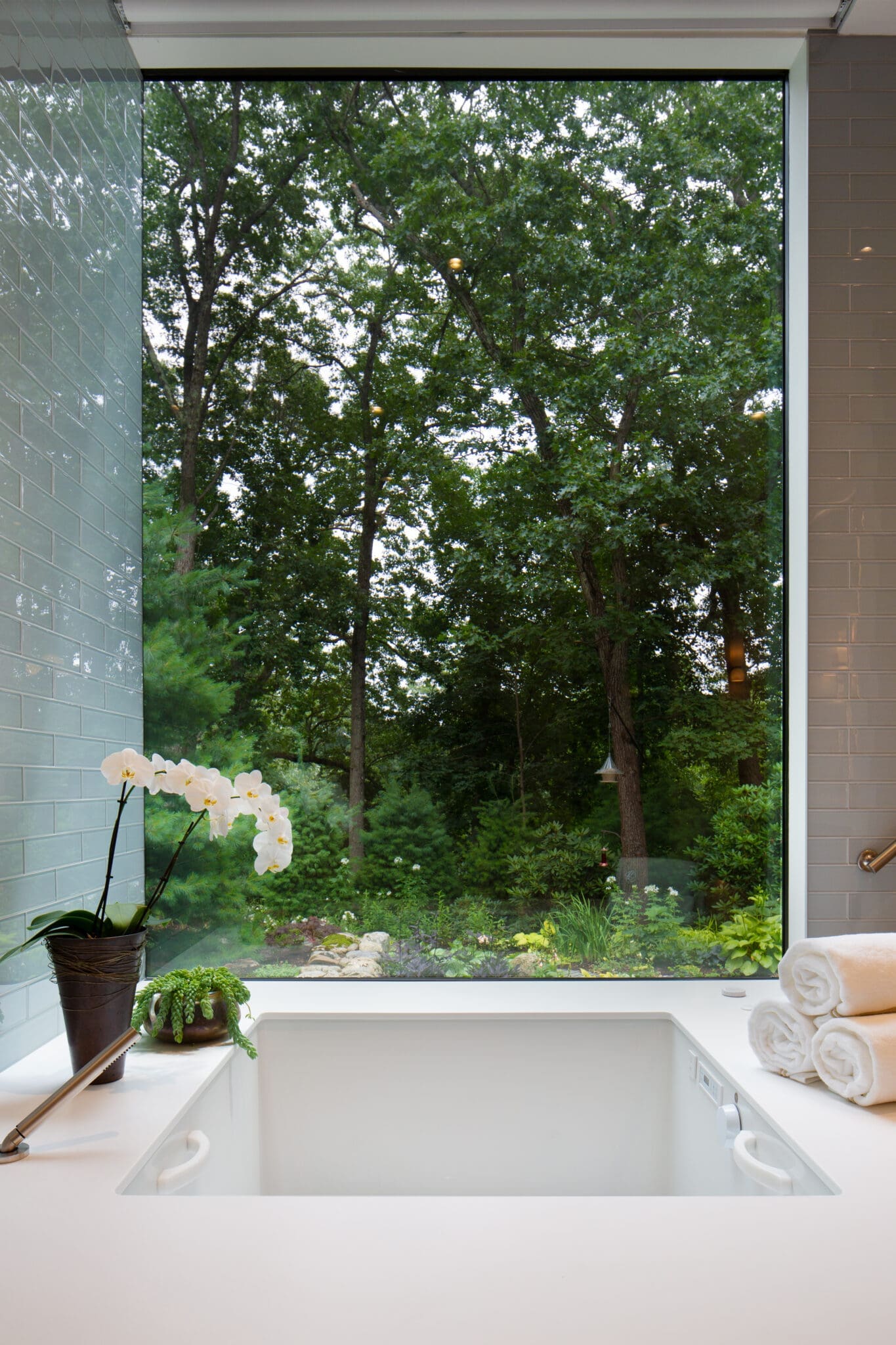 A photo of a deep, square-shaped tub with a thick wrap-around edge. Behind the tub is a large window looking out to the woods.